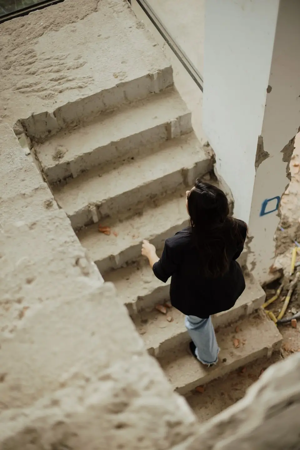 Júlia sobe uma escada de concreto ainda bruto, vista de cima, num canteiro de obra
